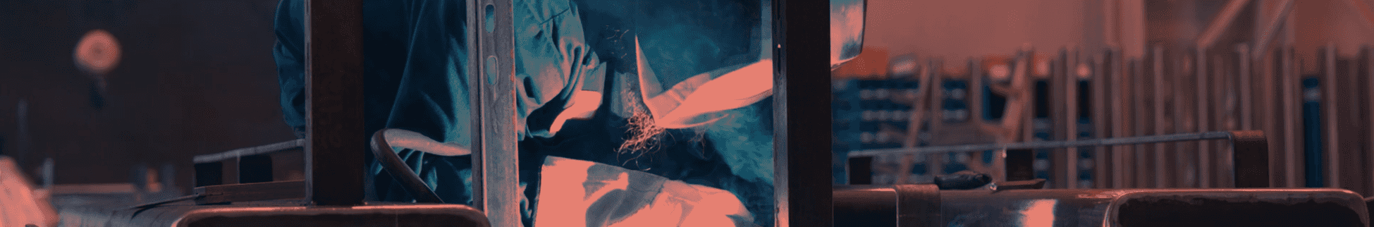 A worker in a blue uniform is welding metal, creating bright orange sparks and blue smoke in a dark industrial setting.
