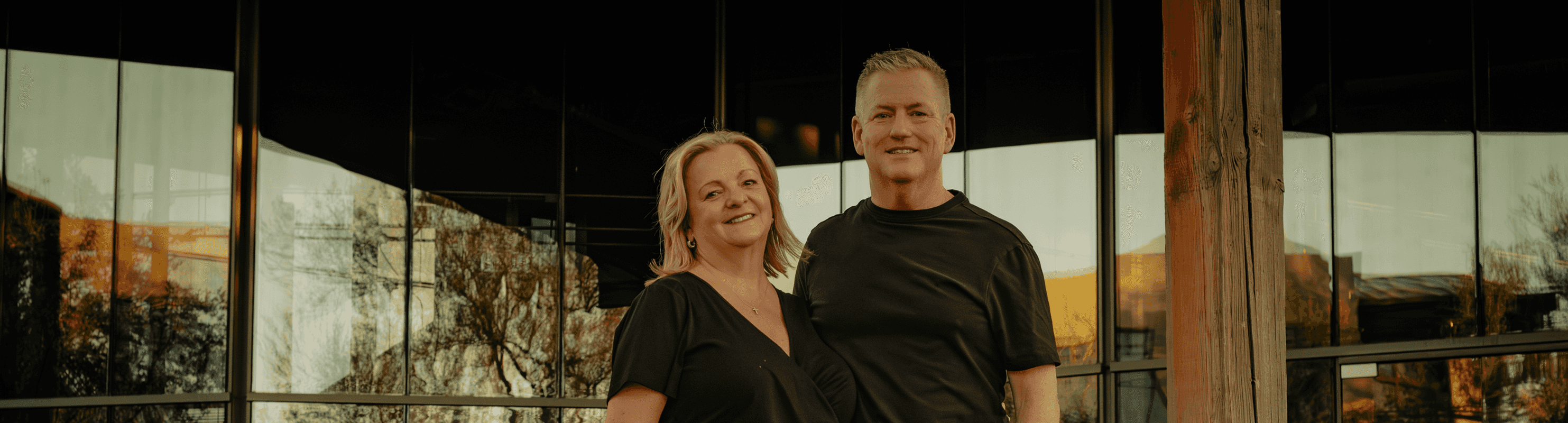 Smiling man and woman standing against a modern building with reflective windows showing trees and other structures.