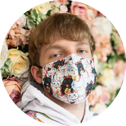 A man with light brown hair and blue eyes wears a white hoodie and a face mask patterned with Maneki-neko lucky cats, surrounded by pastel flowers.