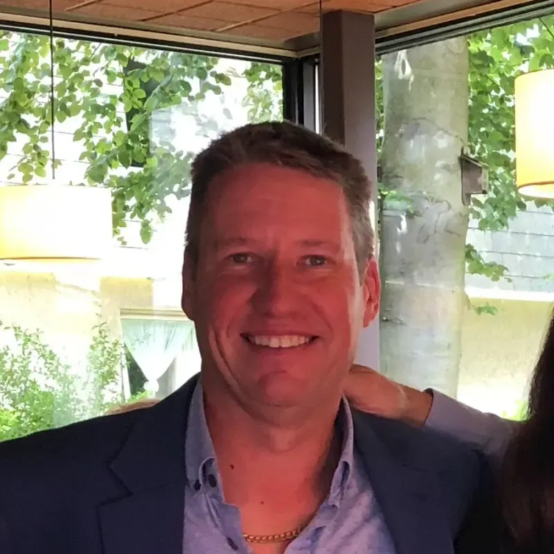 A smiling man in a blue blazer and light blue shirt, wearing a gold chain, stands indoors with a hand on his shoulder and green trees visible outside.