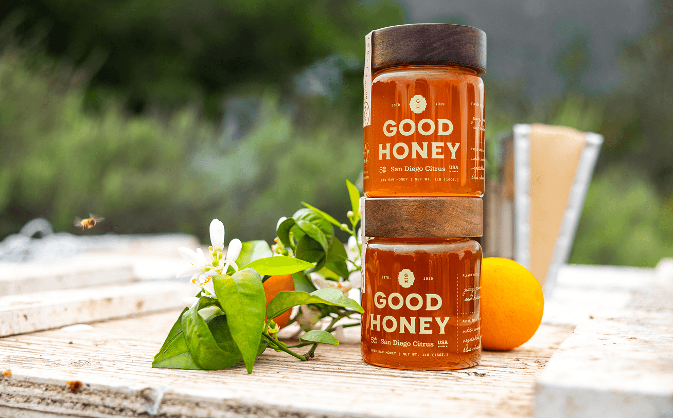 Two stacked jars of Good Honey San Diego Citrus, with an orange, blossoms, and a bee in flight, on a wooden surface near a beehive.