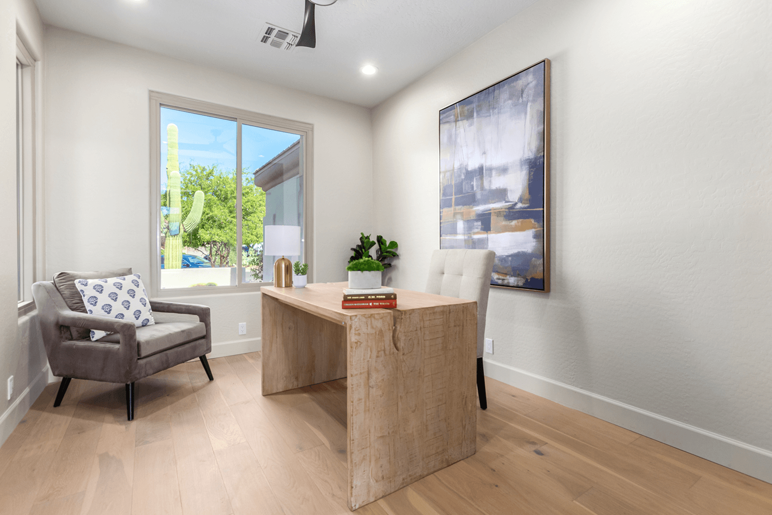 A bright modern home office with a light wooden desk, a grey armchair by a window showing a saguaro cactus, and a large abstract painting.