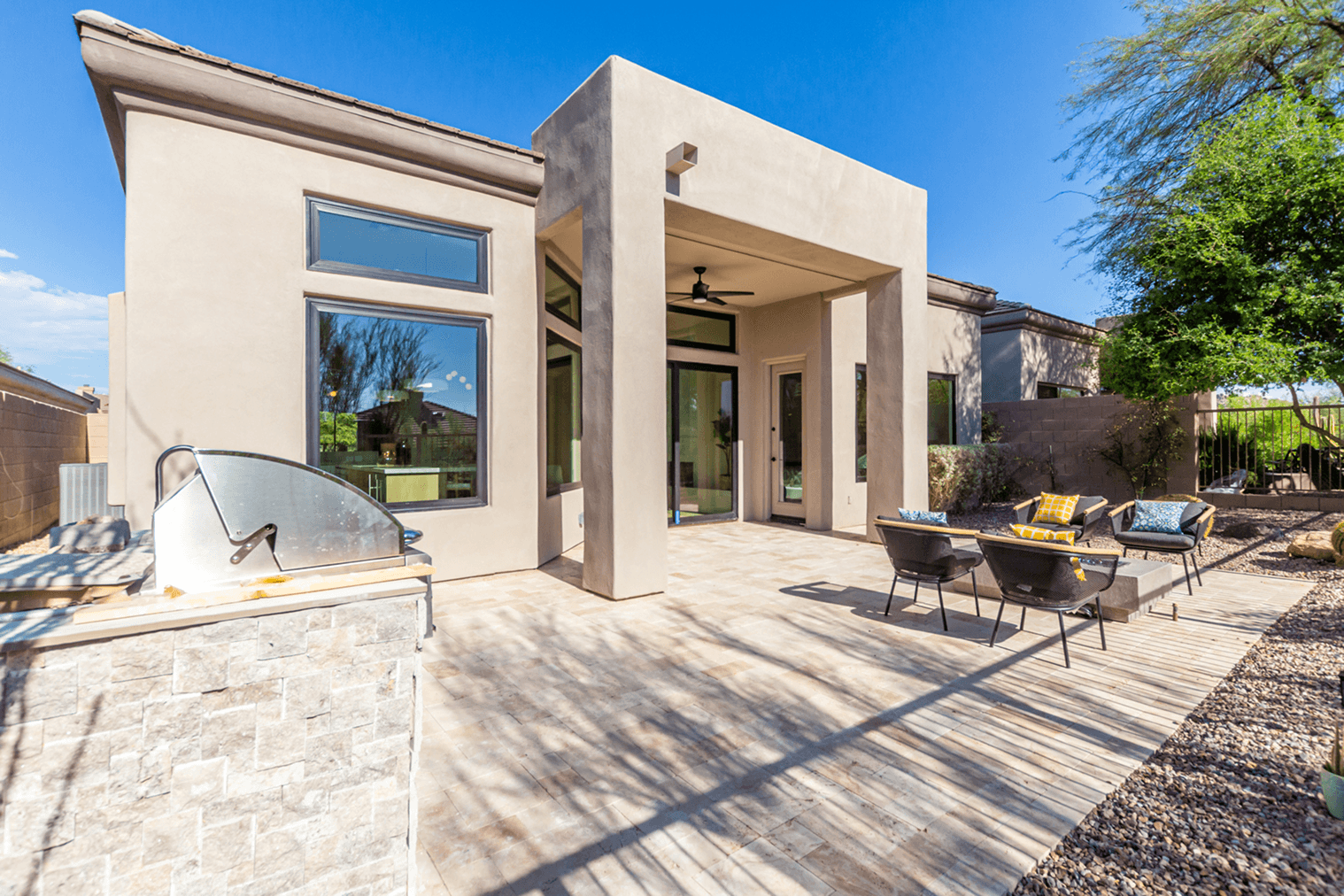 Modern outdoor patio with a built-in grill, a tiled seating area with a fire pit, and a contemporary house under a clear blue sky.