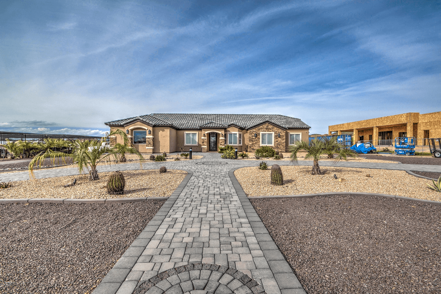 A single-story light brown stucco home with stone accents and a dark tiled roof, featuring a paver walkway and a xeriscaped front yard with palm trees and cacti. A plant nursery is on the left, and a building under construction is on the right.