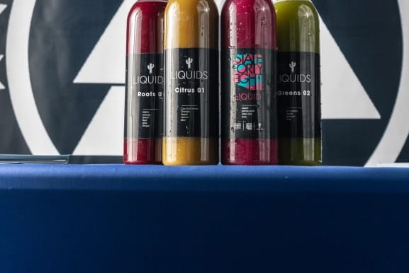 Four cold-pressed juice bottles, in red, orange, magenta, and green, stand on a blue table. Labels include 'Roots 01', 'Citrus 01', 'State Forty Eight Liquid', and 'Greens 02'.