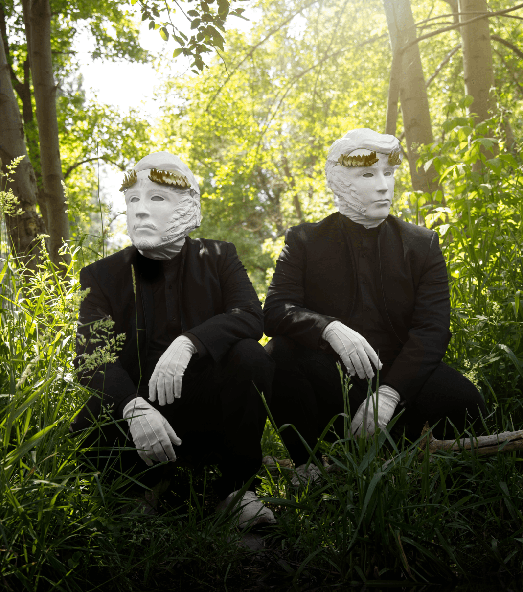 Two figures in black suits, white gloves, and elaborate white masks with golden leaf crowns crouch in a sunlit forest.