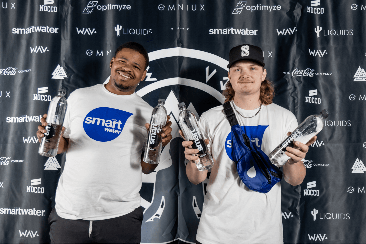Two smiling men hold smartwater bottles in front of a black backdrop featuring logos for Smartwater, NOCCO, Optimyze, Wavy, Liquids, Coca-Cola, and MNIUX.