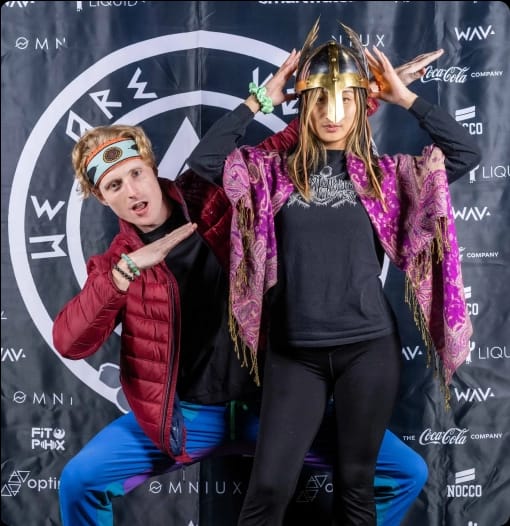 Two people, a man in a red jacket and a woman in a Viking helmet and purple scarf, strike playful poses in front of a branded backdrop.