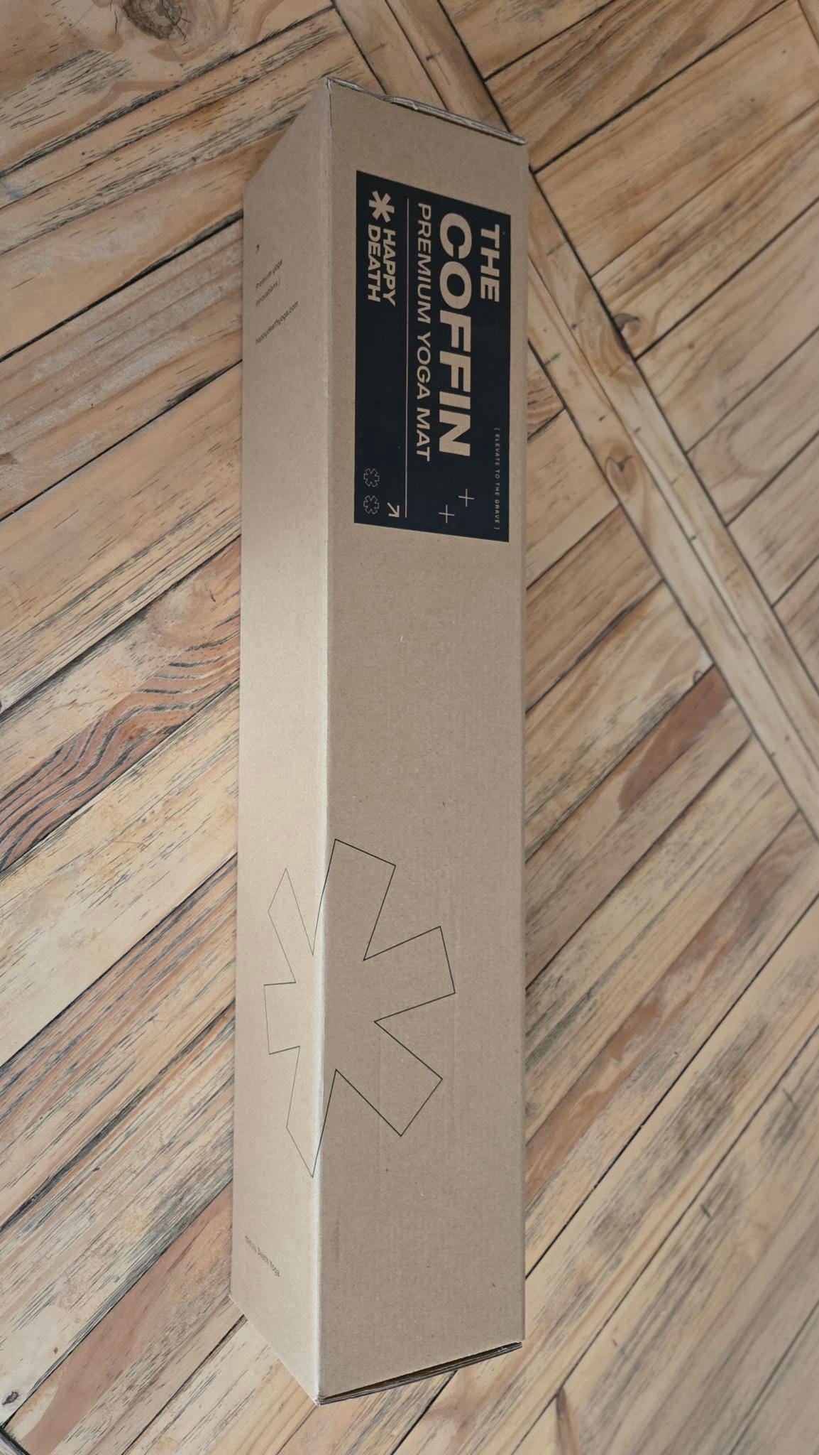 Cardboard box for "THE COFFIN PREMIUM YOGA MAT" by HAPPY DEATH, featuring the tagline "ELEVATE TO THE GRAVE."