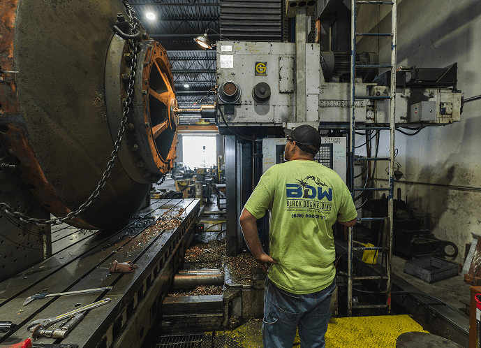 A worker wearing a "Black Dog Welding" shirt observes a massive cylindrical metal part on a machine table next to a large industrial boring mill, surrounded by metal shavings.