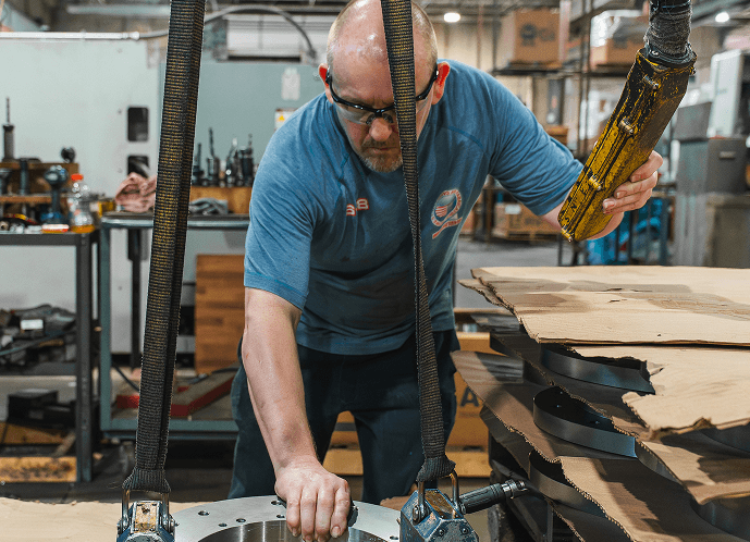 A factory worker in safety glasses positions a large metal flange with his hand while operating a yellow control for an overhead lift.