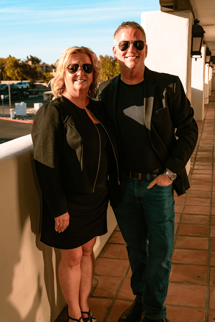 A smiling couple, both in sunglasses, stands on an outdoor balcony next to a white railing. The woman wears a black dress and jacket, and the man wears a black blazer and jeans.