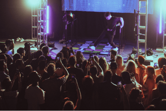 A person plays Dance Dance Revolution on a stage lit by purple and blue lights, while a crowd watches and records with their phones.