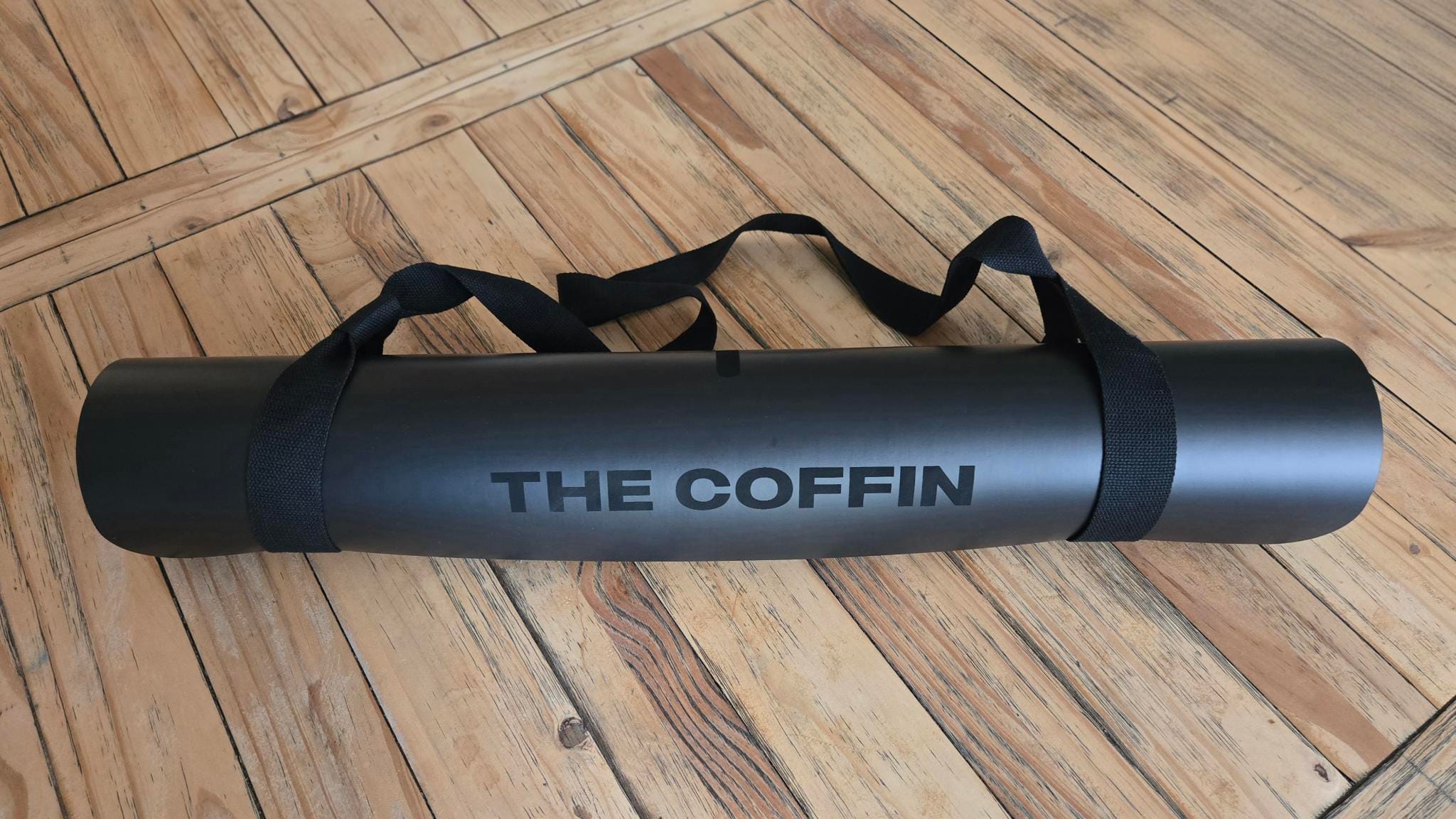 Black cylindrical fitness sandbag with "THE COFFIN" printed on its side and black carrying straps, resting on a wooden floor.