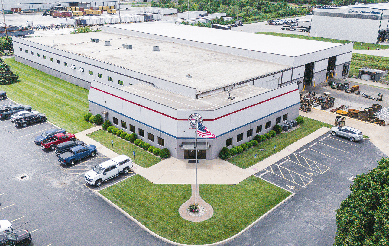 Aerial view of a modern industrial building featuring a front office with red and blue stripes, an American flag, and a company logo, flanked by a parking lot and outdoor material storage.