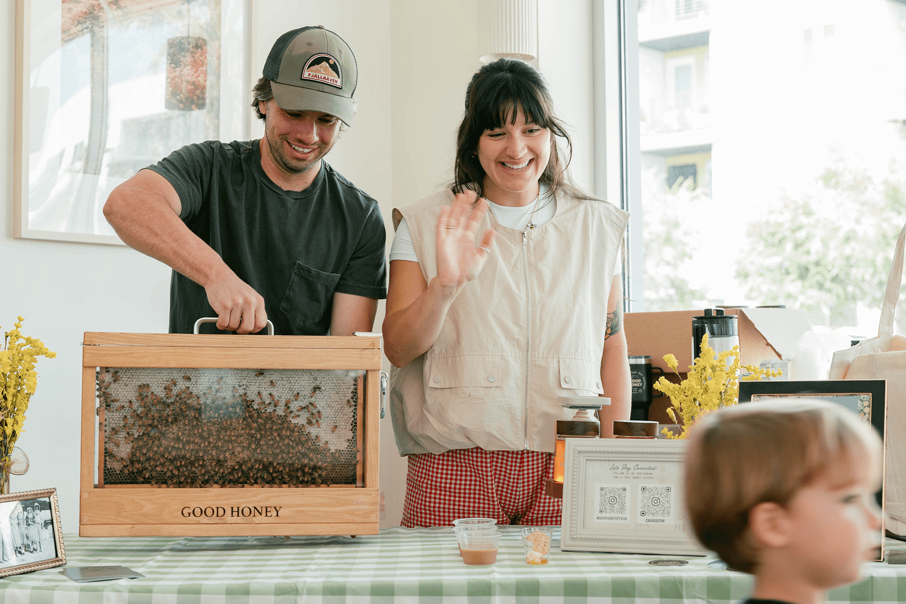Two smiling vendors at a market stall. A man holds a display box of live bees and honeycomb labeled 'GOOD HONEY', while a woman waves, with honey samples on the table.