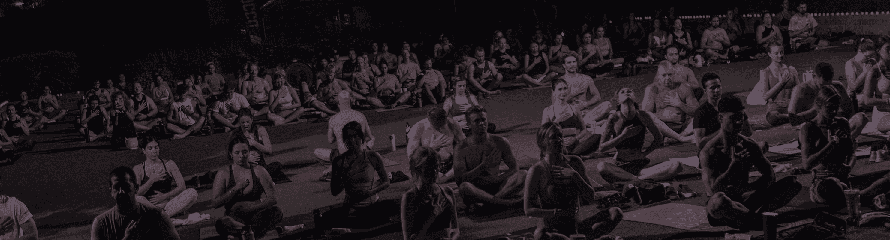 A large group of people sit cross-legged on yoga mats on a grassy area at night, many with hands placed over their hearts in a meditative pose.
